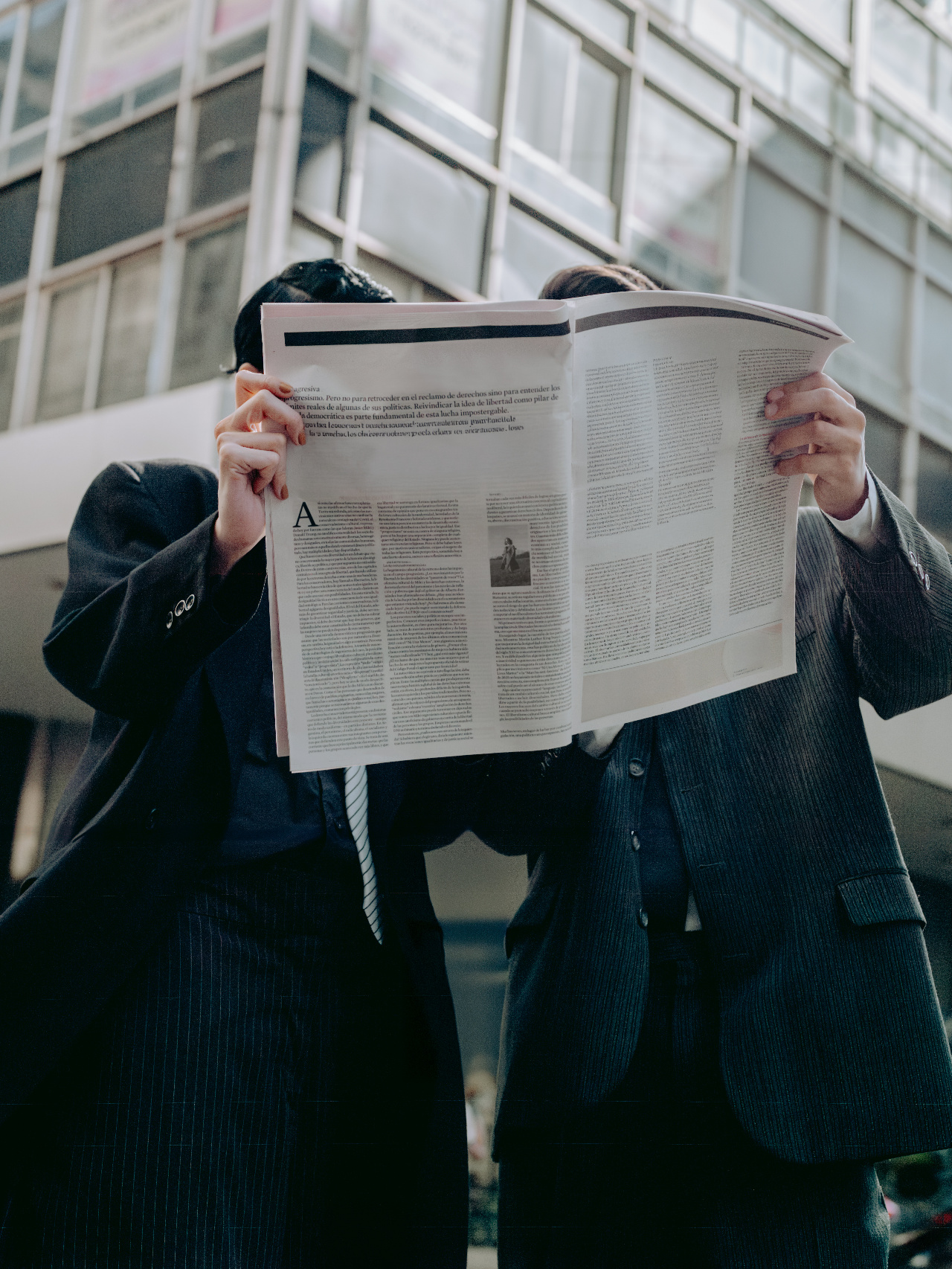 Two professionals in tailored suits holding a newspaper up over their faces