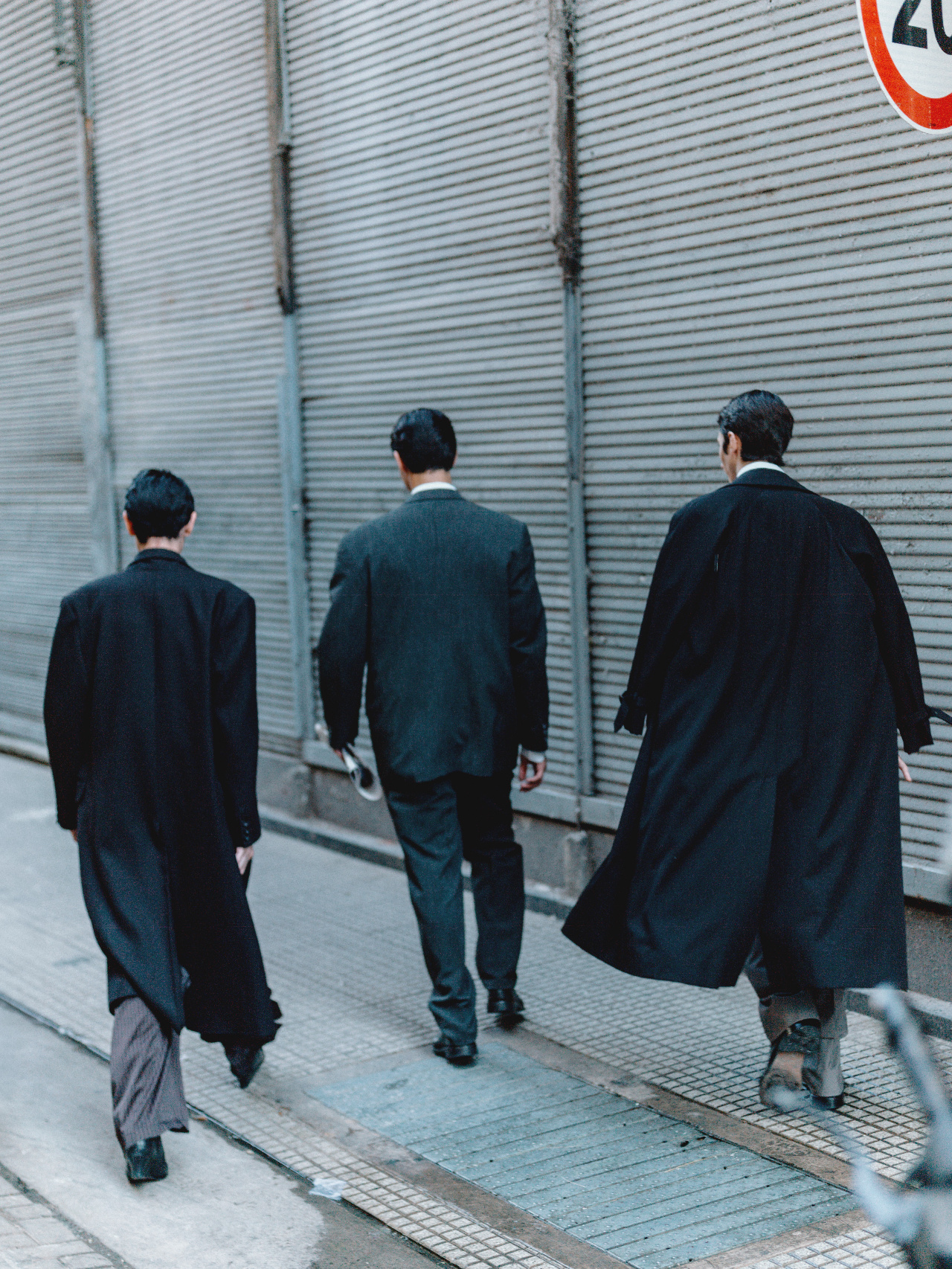 Three figures in tailored coats walking away down a shuttered street